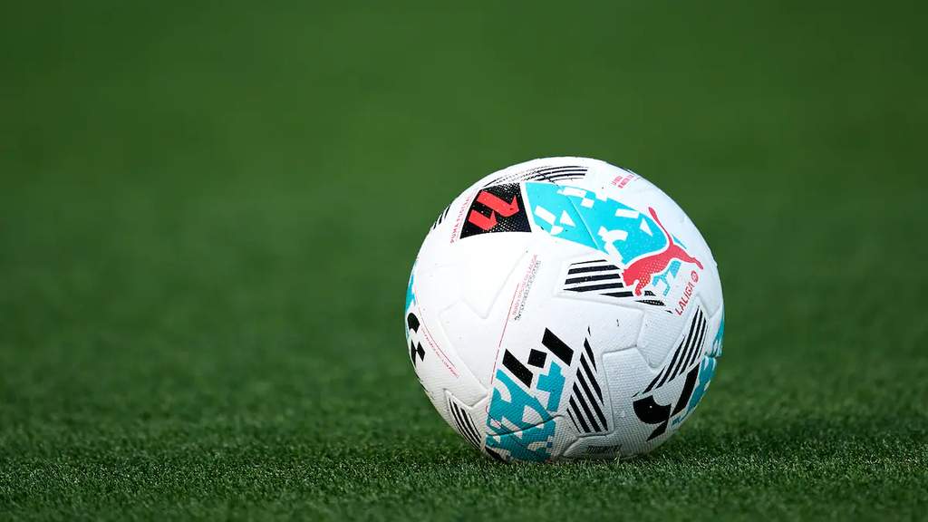 Tampilan aplikasi LaLiga Fantasy di layar ponsel pintar dengan latar belakang rumput hijau stadion sepak bola modern.